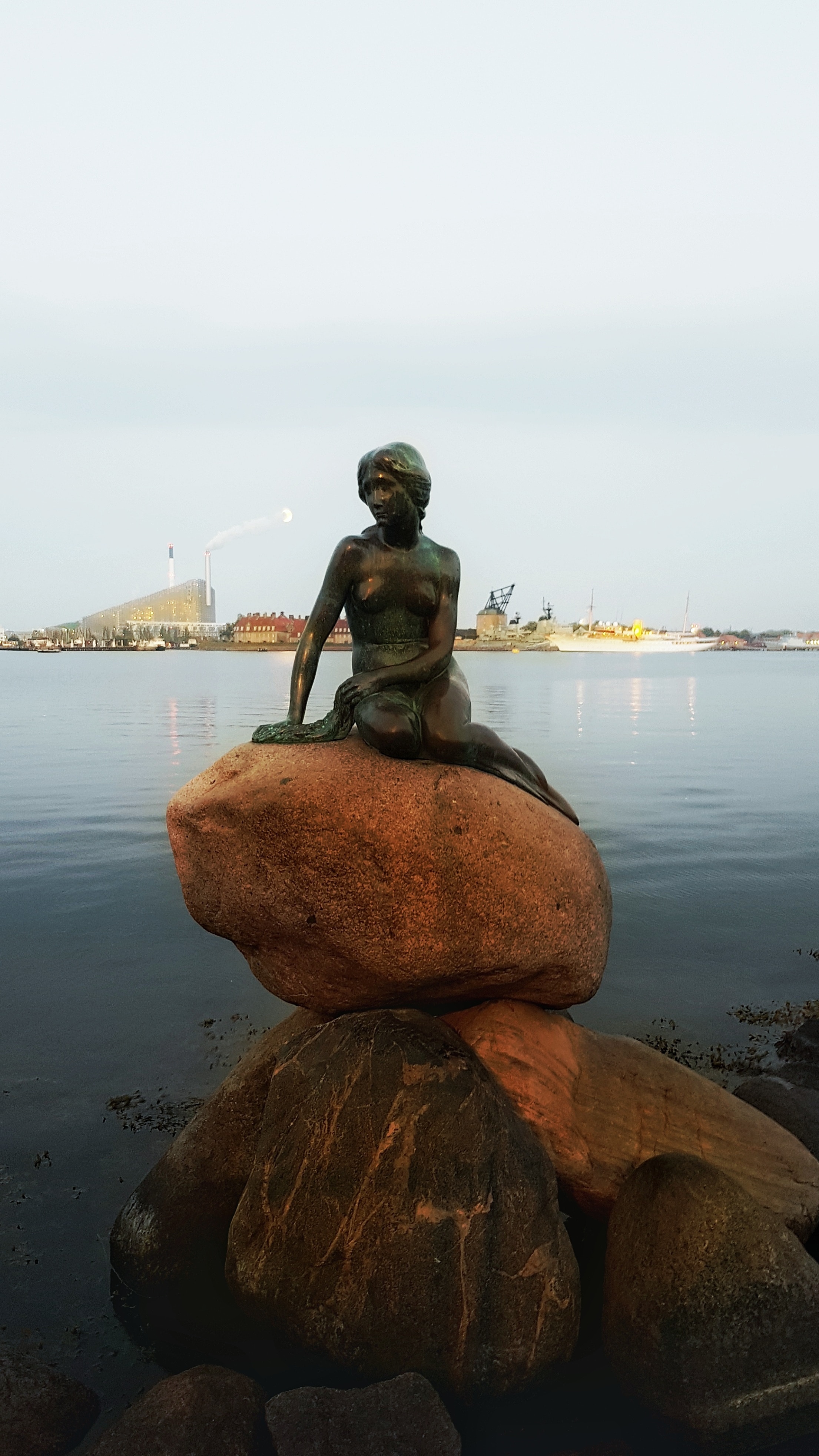 The Little Mermaid sculpture at Langelinie Pier in Copenhagen, Denmark, gazing over calm waters with the city's industrial backdrop and crescent moon.