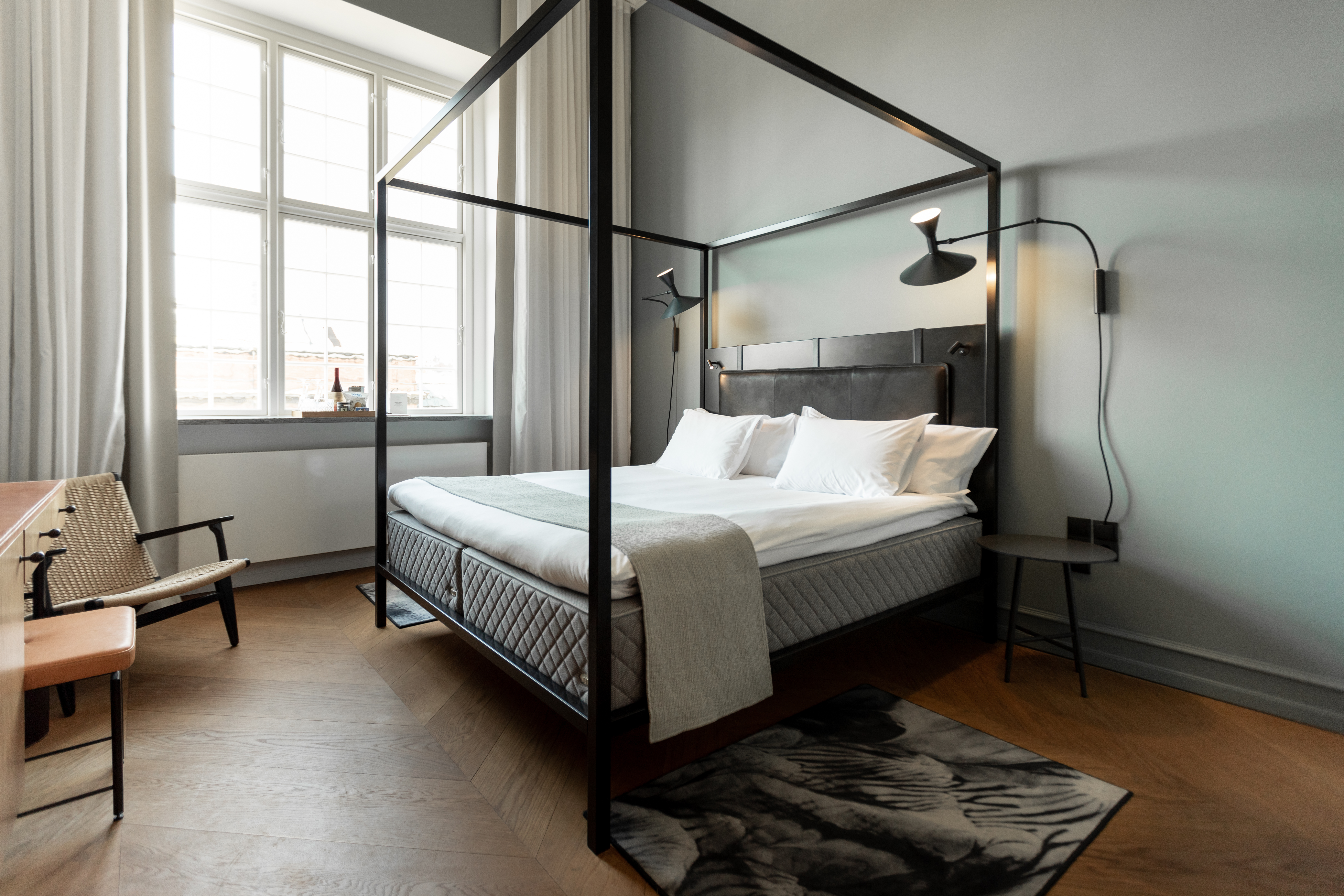 Superior hotel room with high ceilings, hardwood oak floor in chevron pattern, deep window niches, flowing drapes in subtle colors, and a curve-shaped dresser and desk designed by Wingårdhs Architects.