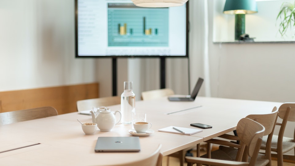 Modern hotel meeting room with projector screen displaying charts, neatly arranged table settings, and contemporary wooden chairs.