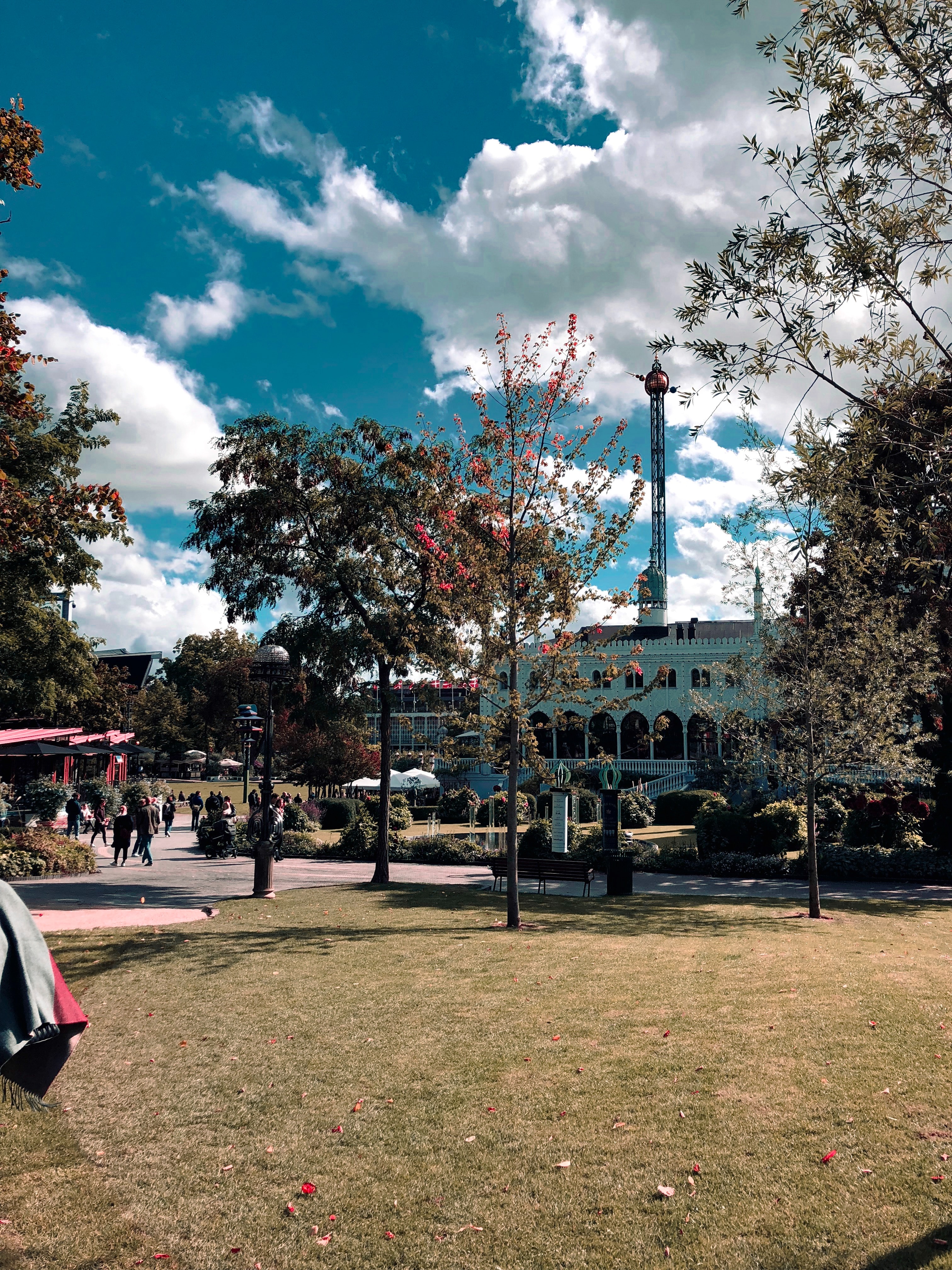 Tivoli Gardens in Copenhagen with visitors walking amidst lush trees, a tall ride tower, and ornate buildings, showcasing the park's blend of amusement and Danish heritage.